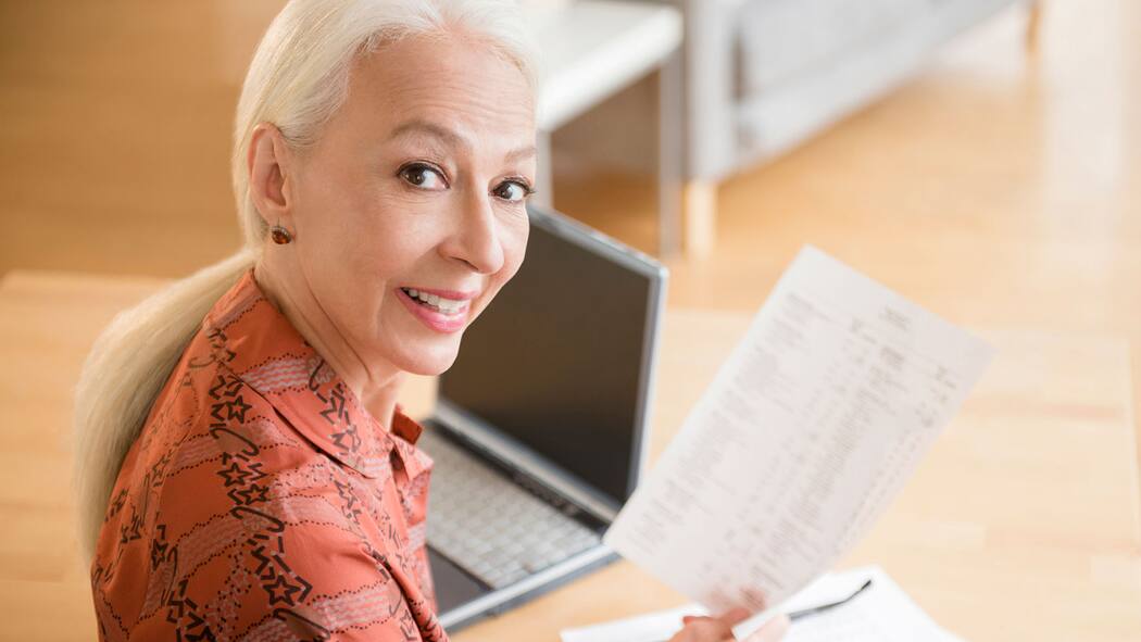 So kommen Sie nicht zu kurz Ältere Frau sitzt an einem Tisch. Auf dem Tisch steht ein Laptop und sie hält einen Auszug in der Hand und schaut in die Kamera.