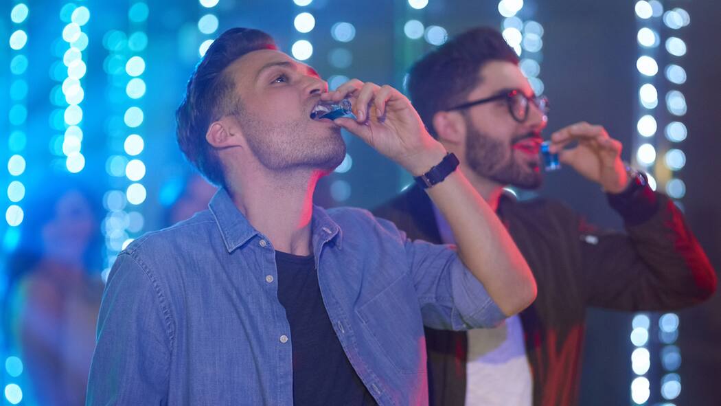 Ist Alkoholsucht vererbbar? Zwei junge Männer trinken einen Shot in einem Club.