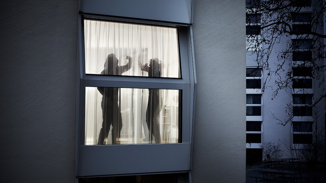 Wann schreitet die Polizei ein? Durch ein Fenster mit Vorhängen sieht man die verschwommenen Konturen eines sich streitenden Paares. Der Mann erhebt seine Hand und will offensichtlich zum Schlag ausholen, während sich die Frau mit den Armen zu schützen versucht.