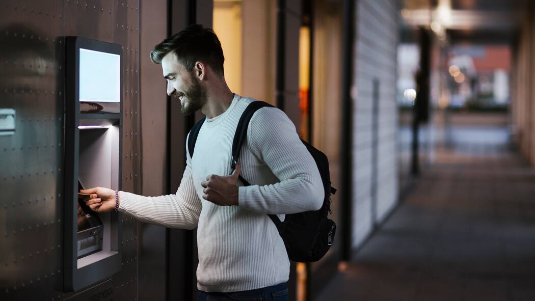 Bleibt mir nur der Bankomat? Ein junger Mann steht vor einem Bankomaten, um Bargeld zu beziehen und schiebt seine Bankkarte in den Automaten.