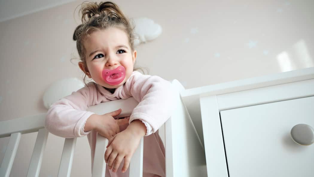 Soll man Kinderwünsche erfüllen? Ein kleines Mädchen steht in ihrem Kinderbett mit Nuggi im Mund und schaut sehnsüchtig nach ihren Eltern.