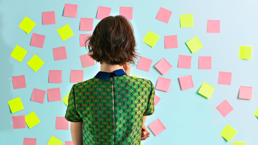 Zeit für einen neuen Job Eine Frau steht mit dem Rücken zur Kamera und blickt auf eine hellblaue Wand, die mit lauter rosa- und gelbfarbigen Post-its beklebt ist.