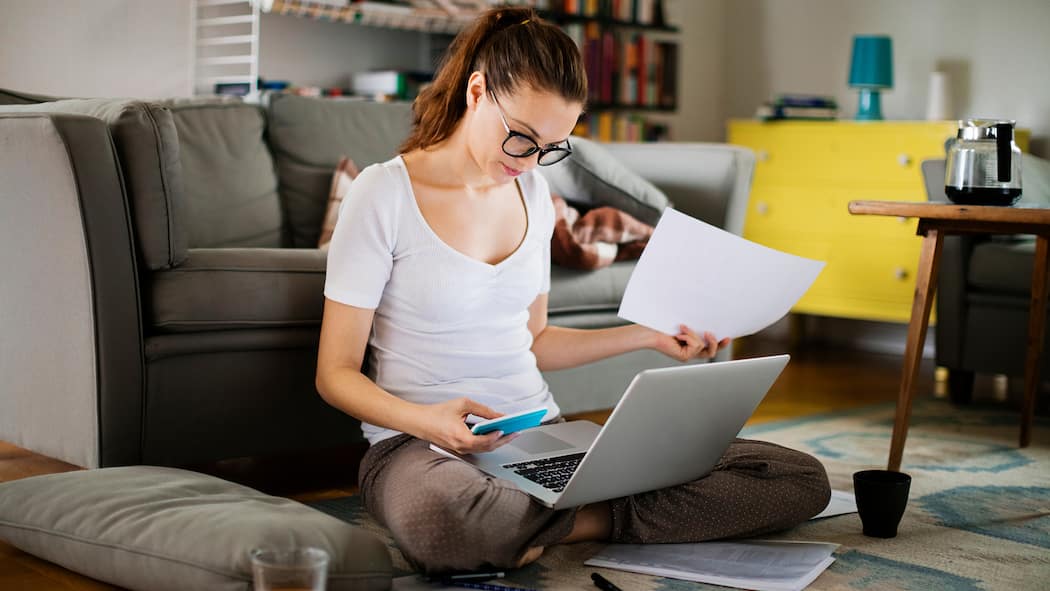 Wann muss ich einzahlen? Eine junge Frau sitzt im Wohnzimmer auf dem Boden und zahlt Rechnungen ein.