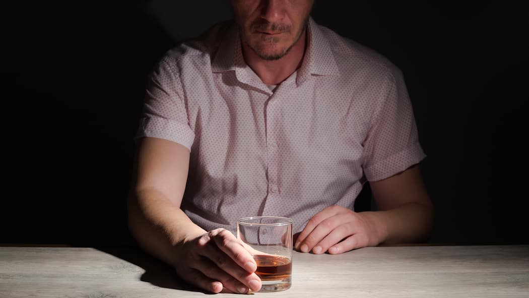 Wenn Genuss zum Zwang wird Ein Mann sitzt im Dunkeln, das Whiskyglas in seiner Hand ist beleuchtet.