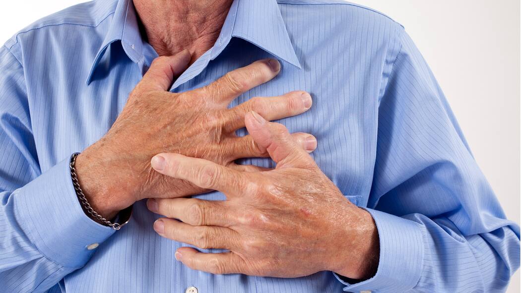 Was fehlt dir, mein Herz? Senior adult man clutching his chest in pain. Heart attack, heart disease. He wears a medical alert bracelet and blue dress shirt.