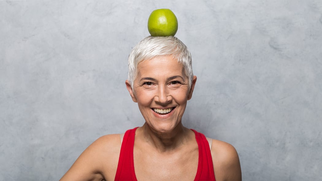 Essen für ein kluges Köpfchen Eine Frau mit grauen Haaren balanciert einen Apfel auf dem Kopf