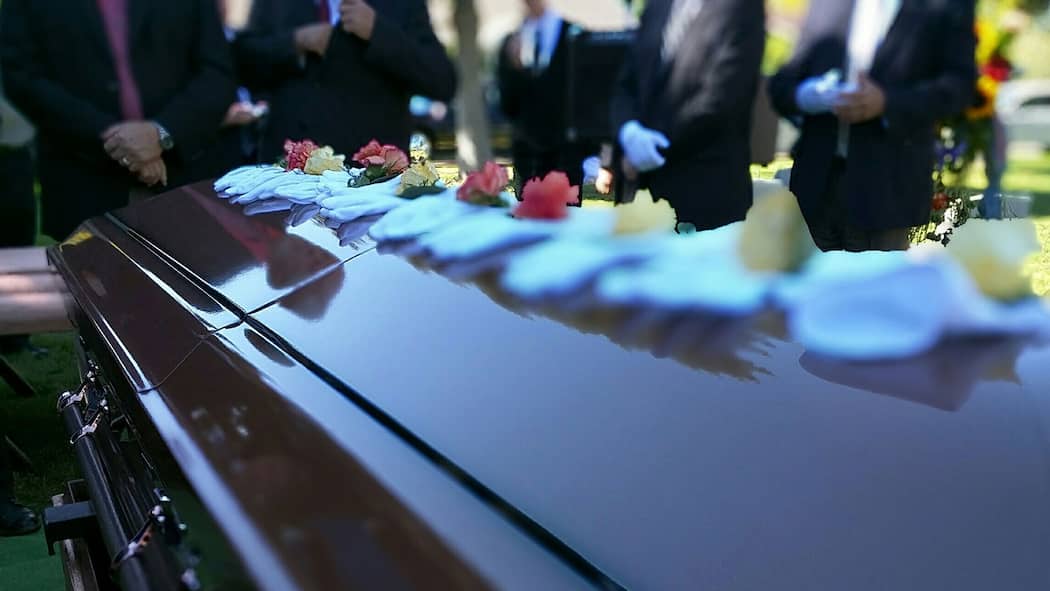 Steuererklärung für Verstorbenen ausfüllen Sarg an Beerdingung auf dem Friedhof und es stehen einige Trauergäste rund herum.