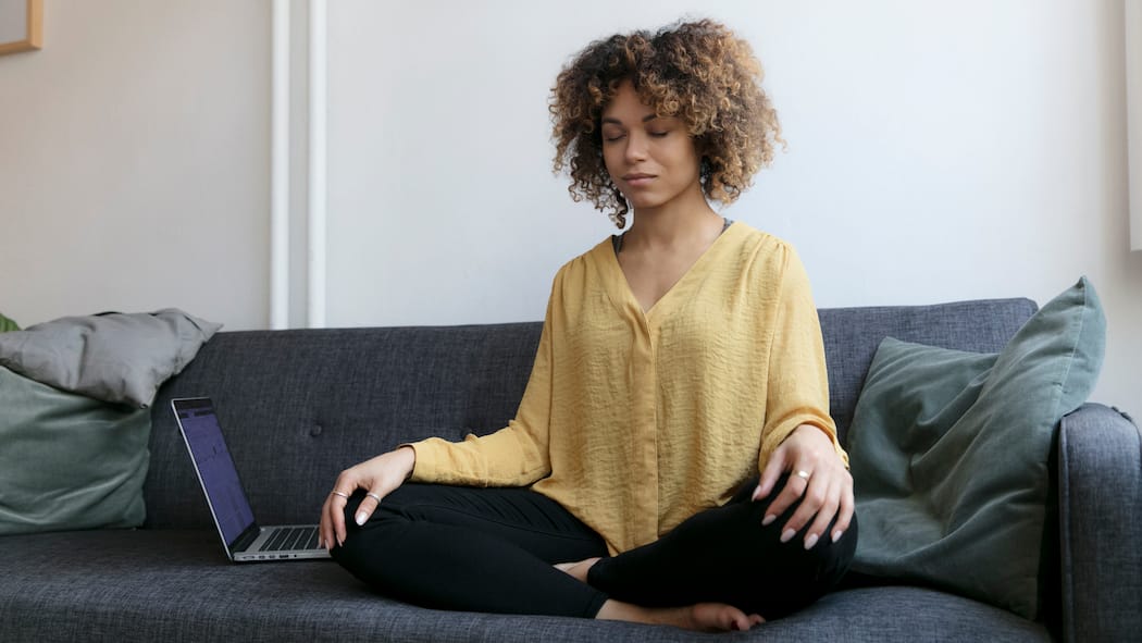 «Raus aus negativen Gedankenspiralen» Junge Frau sitzt mit Laptop auf dem Sofa und meditiert