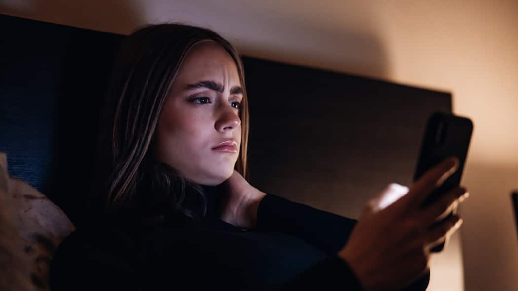 Wie bekommt man Ängste in den Griff? Eine junge Frau liegt mit besorgtem Gesicht im Bett und liest Nachrichten auf ihrem Handy