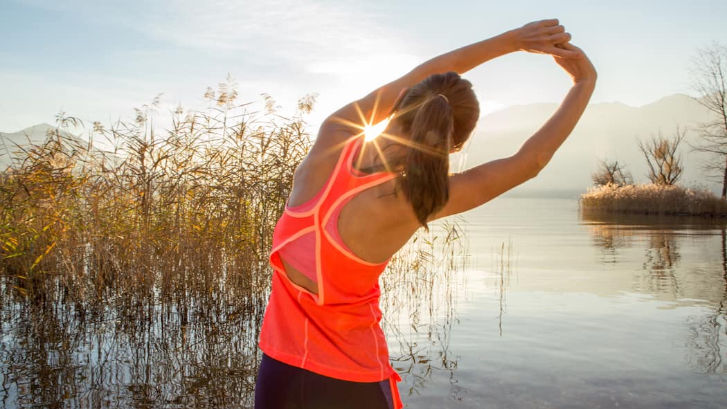 6 Tricks, wie Sport schneller schlank macht Eine junge Frau dehnt sich vor dem Sport draussen in der Natur bei Sonnenaufgang