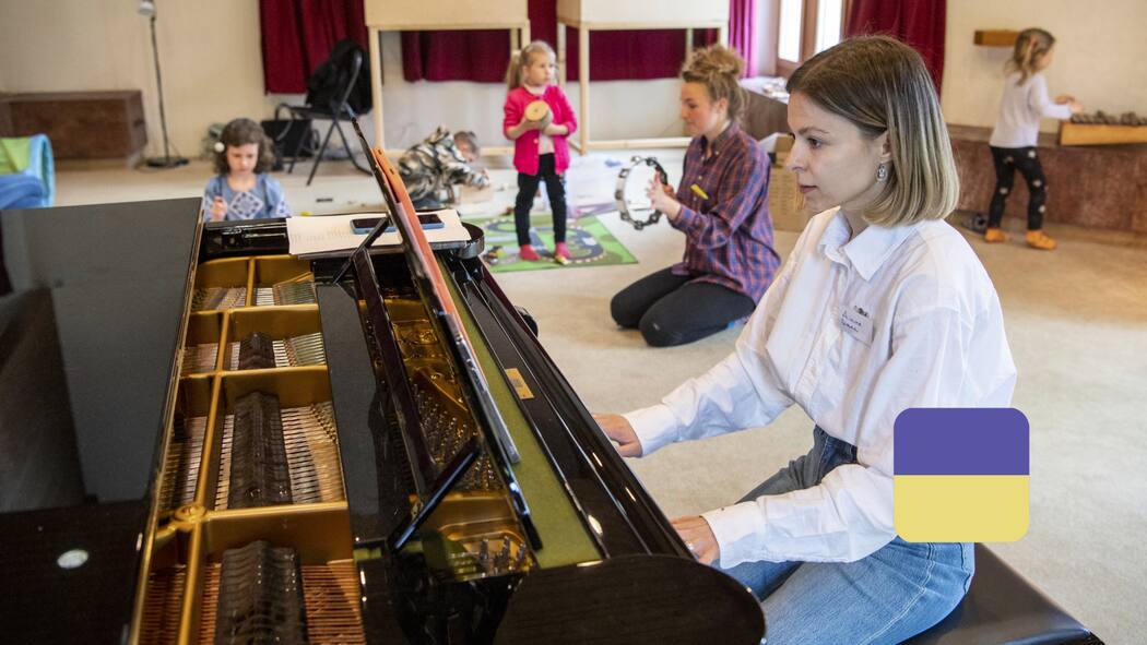 Між експлуататорами та бюрократизмом Eine junge Frau sitzt am Klavier und spielt ein Stück während im Hintergrund kleine Kinder spielen.