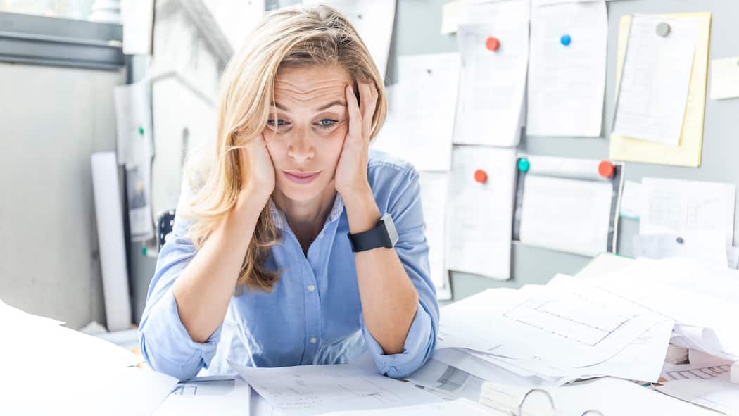 Macht uns die Arbeit krank? Eine Frau sitzt an ihrem Arbeitsplatz, der mit Akten überladen ist, stützt den Kopf in die Hände und macht ein sorgenvolles Gesicht.