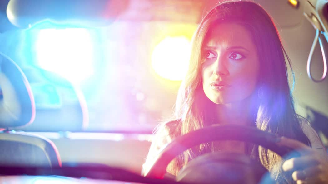 Was sind meine Rechte? Eine junge Frau sitzt am Steuer während hinter ihr die Blaulichtanlage eines Polizeiautos zu sehen ist. Diese Rechte haben Autolenkerinnen und Autolenker, wenn sie von der Polizei kontrolliert werden