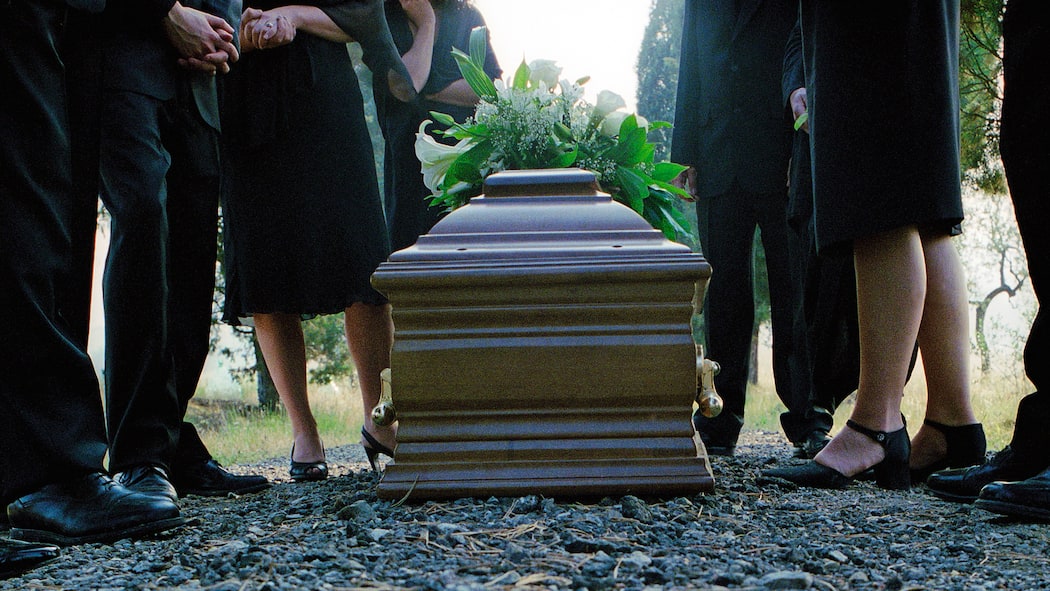 Drei typische Irrtümer Mit Blumen geschmückter Holzsarg an Trauerfeier und Trauergäste stehen um den Sarg herum.
