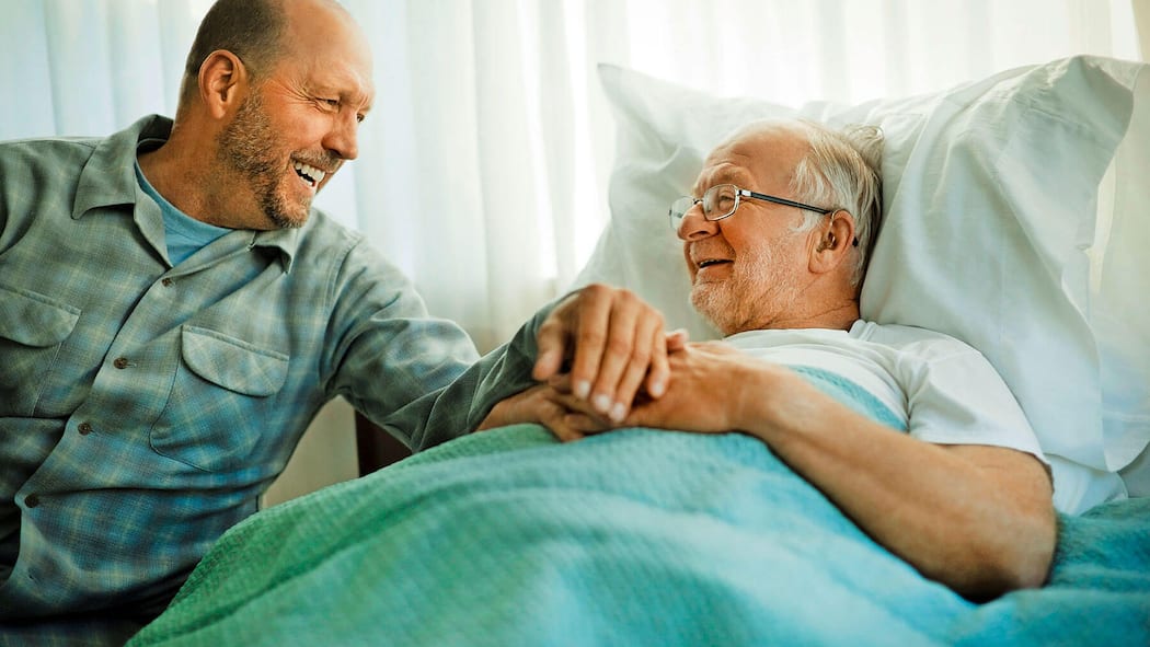 Wozu dient ein Vorsorgeauftrag? Ein älterer Mann liegt in einem Spitalbett und lächelt, ein zweiter Mann sitzt daneben und hält seine Hand. Für den Fall einer Urteilsunfähigkeit gibt es die Möglichkeit, eine Drittperson mit einem Vorsorgeauftrag zu bestimmen.