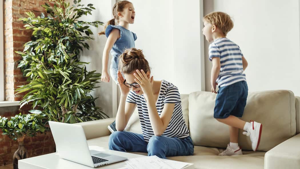 Ein Leben auf der Überholspur Müde junge Mutter sitzt auf dem Sofa und arbeitet am Laptop und an Dokumenten während kleine Kinder Spass haben und Krach machen