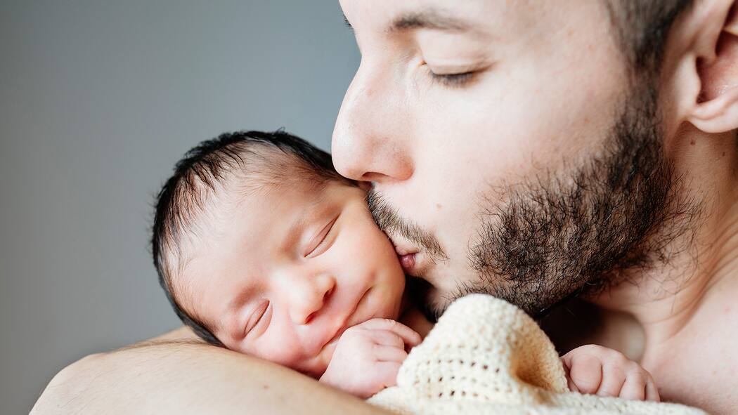 10 Regeln zum Papi-Urlaub Ein frischgebackener Vater hält sein neugeborenes Kind in den Armen und küsst es liebevoll auf die Wange.