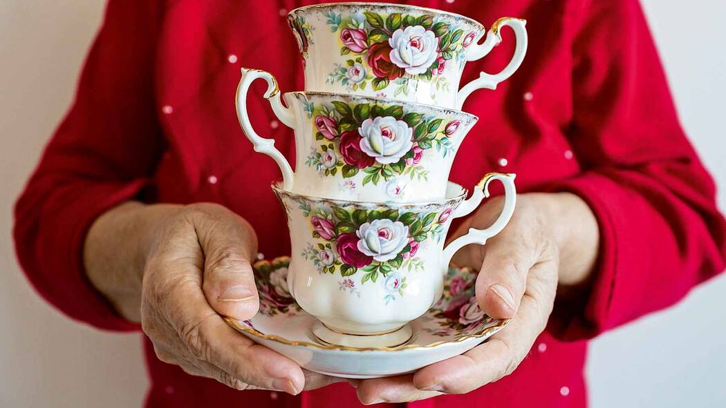 Das Erbe verteilen – oder der Mutter überlassen? Eine ältere Frau hält 3 übereinander gestapelte Teetassen auf einer Untertasse in der Hand. Warum es sinnvoll ist, den gesetzlichen Erbteil zu fordern statt aufs Erbe zu verzichten.