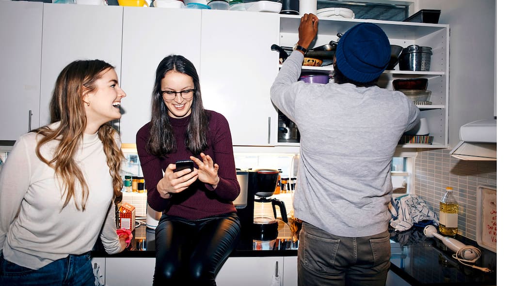 Die erste WG – das sollten Sie beachten Eine Wohngemeinschaft von zwei Frauen und einem Mann befinden sich in einer Küche, in der ein kleines Durcheinander herrscht.