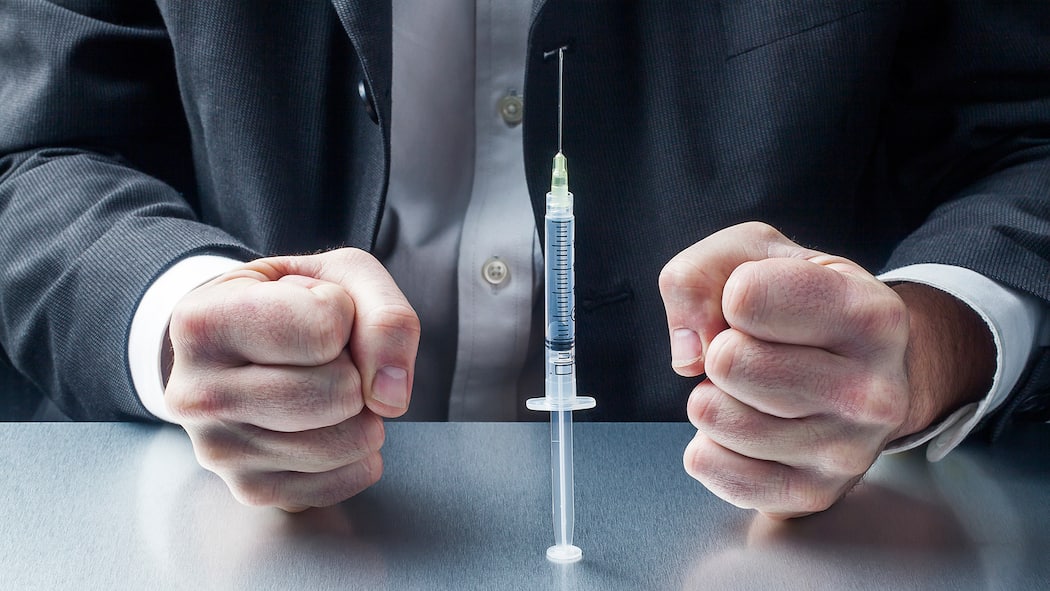 Darf der Chef beim Impfen und Testen mitreden? Ein Mann im Anzug sitzt an einem Tisch, vor ihm steht eine Spritze mit einem Covid-19-Impfstoff. Seine Hände sind zu Fäusten geballt.