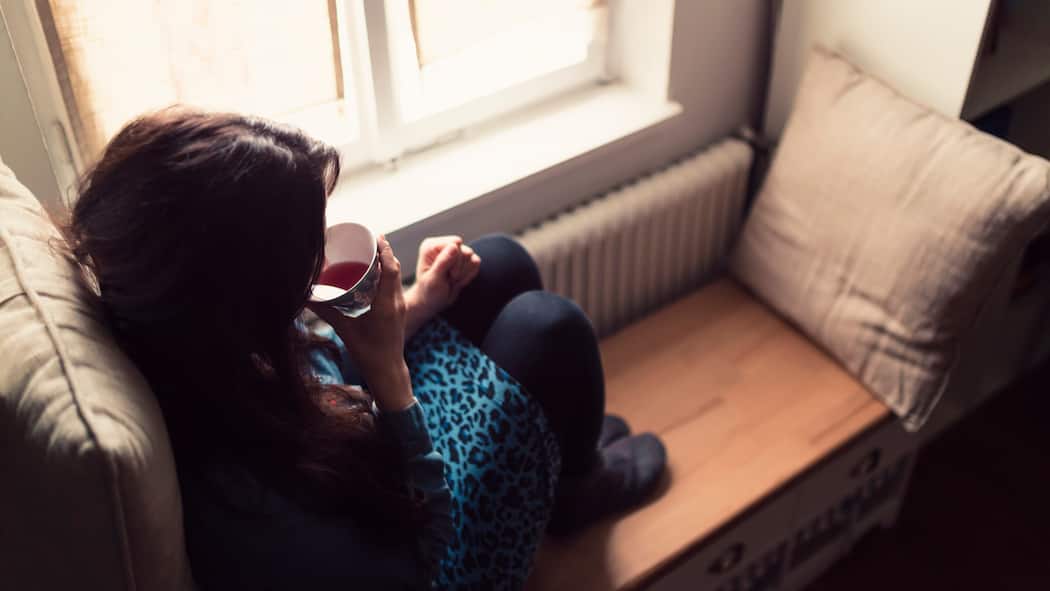 Schlau heizen mit smarten Thermostaten Eine junge Frau sitzt zusammengekauert neben einem Heizkörper und hat sich einen warmen Tee zubereitet.