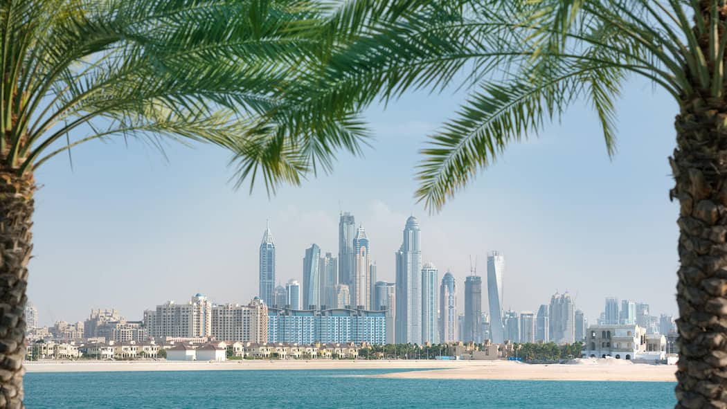 Was kann ich tun, wenn ich nicht mehr nach Dubai will? Skyline der Stadt Dubai von einem Strand aus, der mit Palmen umrahmt ist - Können Pauschalreisende wegen der Reisewarnung des Bundes ihre Reise nach Dubai wegen des Kriegs in Nahost kostenlos absagen? Der Beobachter erklärt, welche Rechte Schweizer Pauschalreisende haben.