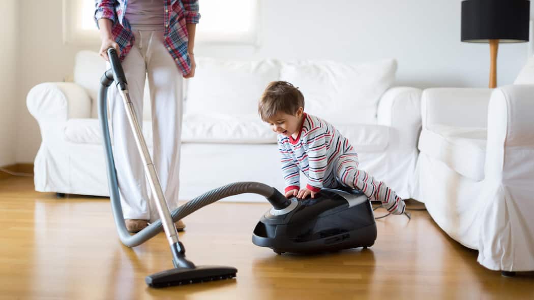 Der Ständerat bremst Frauen aus A mother vacuums the floor of a bright living room while her young child playfully sits on the vacuum cleaner, creating a cheerful atmosphere in their home.