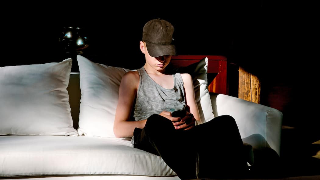 Dürfen Eltern bei Verdacht rumstöbern? Ein minderjähriger Junge sitzt auf dem Sofa in einem verdunkelten Zimmer und sieht sich heimlich Bilder auf seinem Handy an. Wenn es um den Besitz von Nacktbildern Minderjähriger geht, kennt das Gesetz kein Pardon. Dennoch sollten Eltern bei einem Verdacht nicht einfach das Handy kontrollieren.