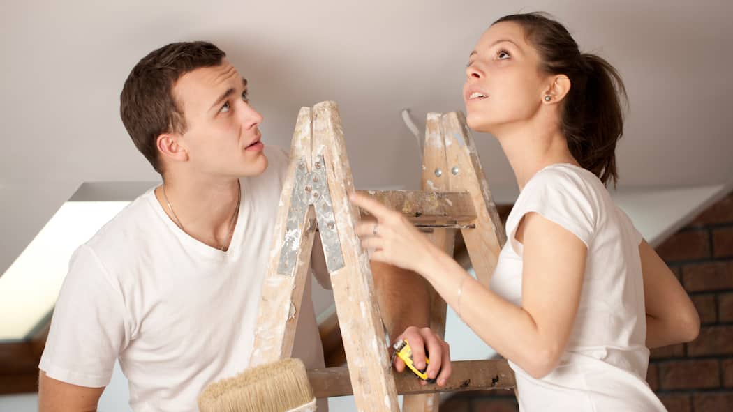 Welche Schäden müssen Mieter zahlen? Junges Paar steht auf einer Leiter und trifft Vorbereitungen, um anschliessend die Decke neu anzustreichen