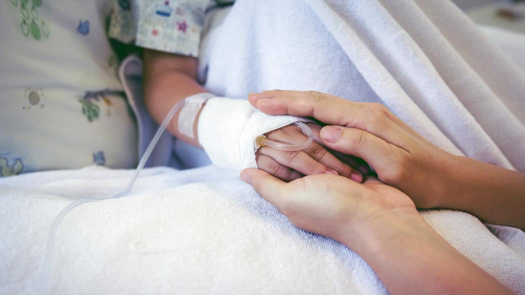 Bekommt man frei, wenn das Kind krank ist? Ein kleines Kind liegt in einem Spitalbett, die Mutter hält ihre Hand in ihren.