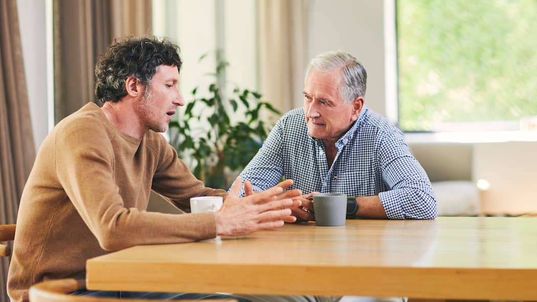 Ist ein Darlehen der Eltern das Beste? Ein erwachsener Sohn und sein Vater sitzen am Tisch währen der Sohn mit den Händen gestikulierend seine finanzielle Situation dem Vater schildert.