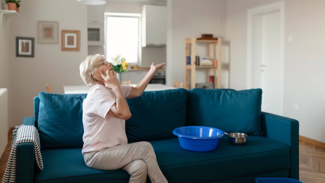 Wann gibts Geld vom Vermieter? Ältere Dame sitzt auf dem Sofa. Auf dem Sofa liegen Eimer. Sie telefoniert und schaut an die Decke hoch.