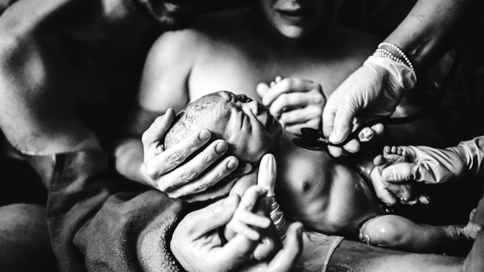 Mutter hält frischgeborenes Kind, dessen Herzschlag gerade ärztlich untersucht wird