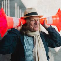 ... was macht eigentlich ein Klanggärtner? Andres Bosshard hält sich zwei orange Warnkegel an die Ohren wie alte Hörgeräte - Andres Bosshard sieht mit den Ohren. Der 70-jährige Musiker veranstaltet Klangspaziergänge und berät Behörden. Sein Lieblingsgeräusch? Menschengemurmel