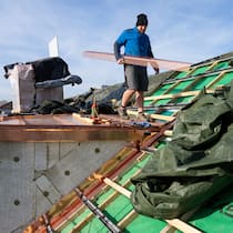 Dieser Fehler kommt Sie teuer zu stehen Ein Mann trägt einen Balken auf dem abgedeckten Dach eines Hauses. Darauf sollten Sie Ihr Hausdach vor Installation einer Fotovoltaikanlage prüfen.
