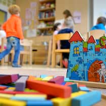 Kita stellt Lernende kurz vor Abschluss frei In einer Kita liegen Spielsachen am Boden, dahinter spielen Kinder und sitzen an Tischen.