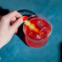 K.-o.-Tropfen im Glas: Wie schütze ich mich? eine manikürte Hand rührt in einem tiefen Cocktailglas, das eine knallrote Flüssigkeit enthält.