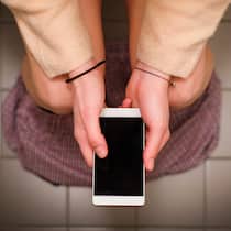WC-Pausen bei der Arbeit – was gilt? Aus der Perspektive einer Person, die mit heruntergelassenen Hosen auf dem WC sitzt und ihr Handy in der Hand hält.