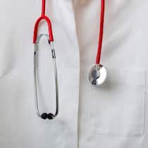 Hausarzt gewechselt – und plötzlich steigt die Prämie Doctor's coat hanging on a coat hanger on a coat rack, with a stethoscope