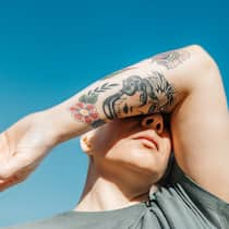 Weg mit dem Tattoo – bloss wie? Frau hält sich tätowierten Arm vors Gesicht - Nicht jede Tätowierung passt ein Leben lang. Doch die Zeichnung weglasern zu lassen, ist nicht nur teuer und langwierig – es ist auch mit Risiken verbunden