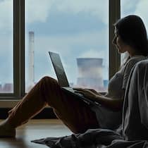 Wie höflich muss man zu einer KI sein? Silhouette einer Frau, die vor einer Fensterfront am Boden sitzt und am Laptop arbeitet