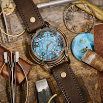 Muss ich das schmucke Stück versteuern? Eine Uhr mit braunem Lederarmband und türkis-farbigen Zifferblatt liegt auf einem Holztisch mit Utensilien eines Uhrmachers.