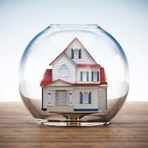 Wird meine Hypothek teurer, wenn die Immobilienblase platzt? Ein Haus steht auf einer hölzernen Oberfläche und wird von einem leeren Fischglas bedeckt, was einer Blase ähnlich sieht. Überbewertete Immobilienpreise könnten ebenfalls wie eine Blase platzen, sollten die Preise markant fallen.