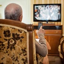 Im Heim und trotzdem TV-Gebühren zahlen? Ein älterer Herr sitzt in einem Sessel und bedient mit der Fernbedienung ein TV-Gerät.