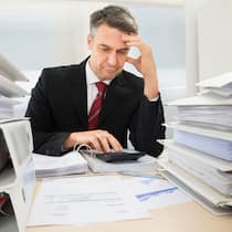 Wenn kein Arbeitslosengeld mehr kommt  Mann im Anzug sitzt in Mitten einer Menge gestapelten Ordnern und rechnet auf dem Taschenrechner etwas aus.