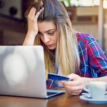 Checken vor dem Klicken Eine junge Frau sitzt angestrengt vor ihrem Laptop, stützt mit der rechten Hand ihren Kopf ab und hält in der linken Hand eine Kreditkarte.