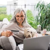 Soll ich auf E-Banking umsteigen? Eine ältere Frau sitzt in ihrem Wohnzimmer auf dem Sofa, ihr Hund auf dem Schoss. Ihr Blick ist auf ein Laptopbildschirm gerichtet, während sie in der anderen Hand eine Bankkarte hält.