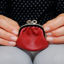 Wie sollen sich Kinder an den Haushaltskosten beteiligen? Eine junge Frau hält ein kleines rotes Portemonnaie, das auf ihrem Schoss liegt, mit den Fingerspitzen fest. Die Frage, wie viel Jugendliche von ihrem Lehrlingslohn zu Hause abtreten sollen, hängt ganz vom Lohn ab und ist abhängig vom Einzelfall. Das sollten Eltern dazu wissen.