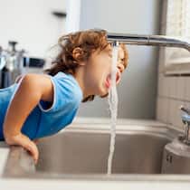 Woher kommt es? Wer überprüft die Qualität? Ein kleiner Junge lehnt sich über ein Spülbecken und streckt seinen Kopf zum Trinken unter den Wasserstrahl. Woher kommt eigentlich das Schweizer Leitungswasser und welche Anforderungen muss es erfüllen, damit wir es problemlos trinken können?