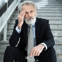 Mit 61 entlassen, weil die Motivation fehlte Ein älterer Mitarbeiter in dunklem Anzug und Krawatte sitzt nachdenklich auf einer Treppe.
